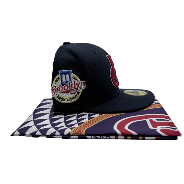 A navy blue Brooklyn Bodega fitted cap resting on a folded BBA scarf made to accompany. The cap features a gold New Era 59FIFTY sticker on the brim and embroidered front and side logos.