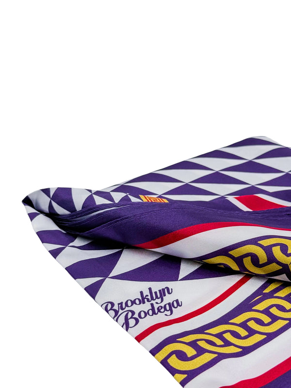 A folded silk scarf featuring a geometric pattern of purple, white, and red triangles, with a yellow Greek key border and 'Brooklyn Bodega' text printed in purple.